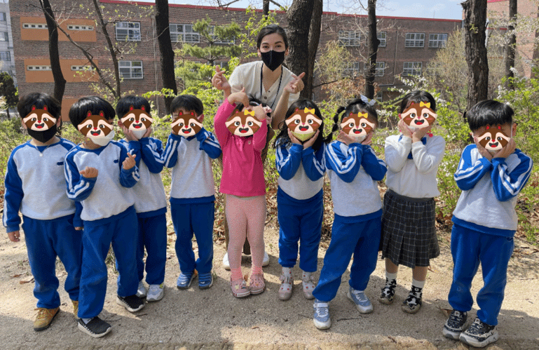 an English teacher and group of Korean kindergarten students posing outside with trees in the background in front of i-garten institute