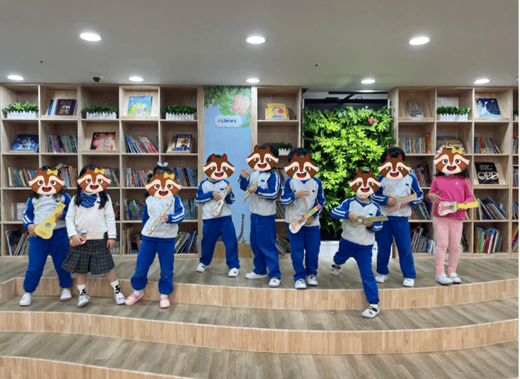 nine Korean kindergarten students holding fake instruments and posing on a small stage in front of a library at i-garten institute