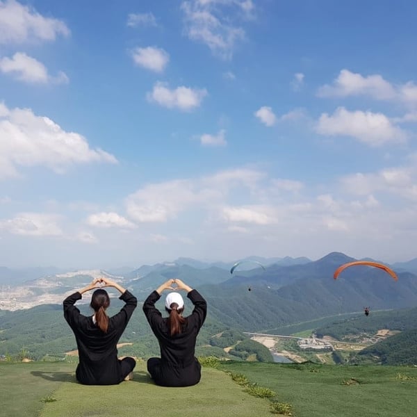 two women sitting watching paragliders with their hands in a heart pose