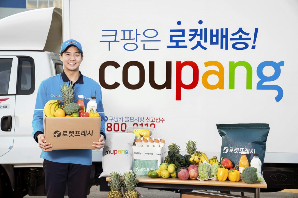 Delivery man holding groceries in front of a delivery truck for Coupang, a Korean grocery delivery service.